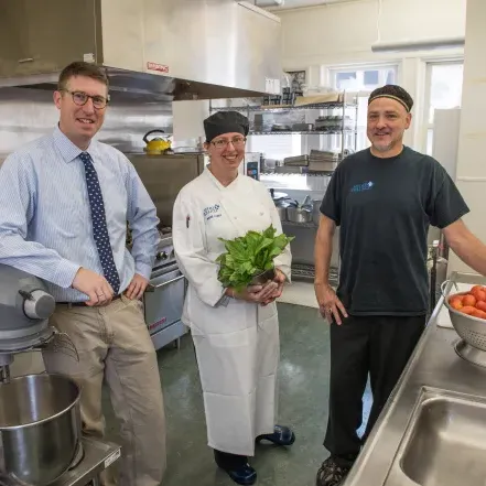 Three members of Dining staff in a kitchen. One man is near a bowl of tomatoes, and a woman is holding a bundle of greens.